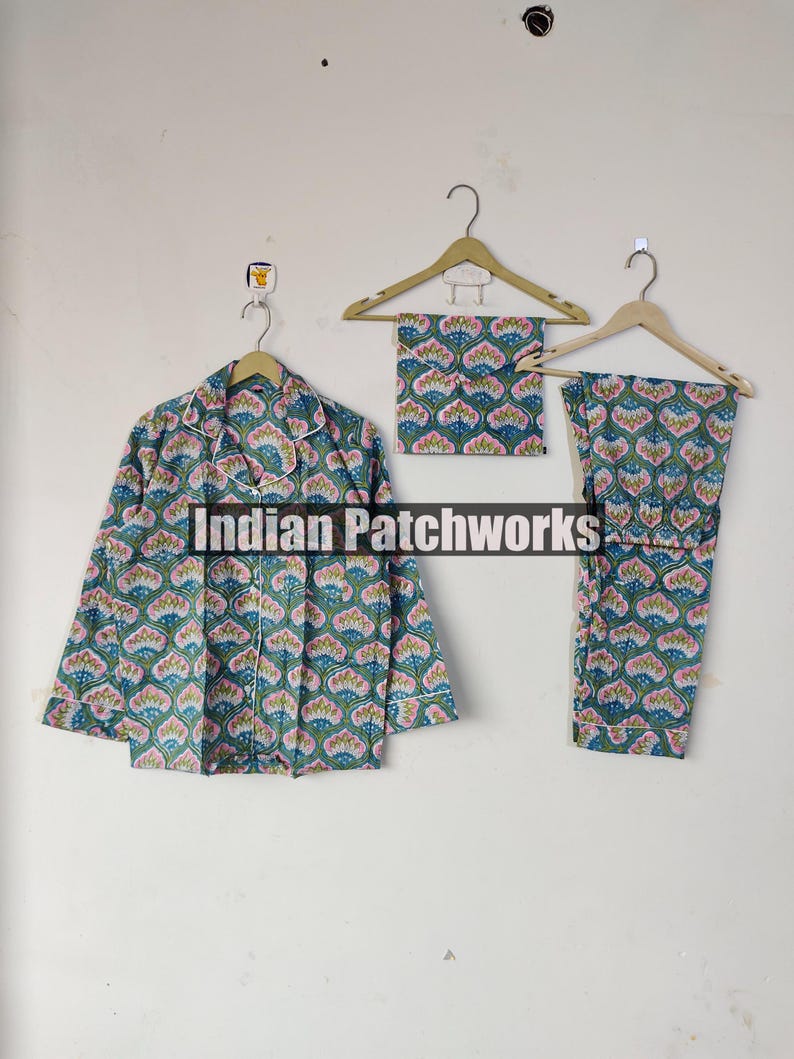 May include: A three-piece pajama set with a floral pattern in shades of green, pink, and blue. The set includes a button-down shirt, a pair of pants, and a matching eye mask, all hanging on wooden hangers. The text "Indian Patchworks" is visible.