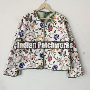 Op de afbeelding: Een wit gewatteerd jack met een bloemenprint en groene bies. Het jack heeft een knoopsluiting en lange mouwen. De tekst "Indian Patchworks" is zichtbaar op de voorkant van het jack.