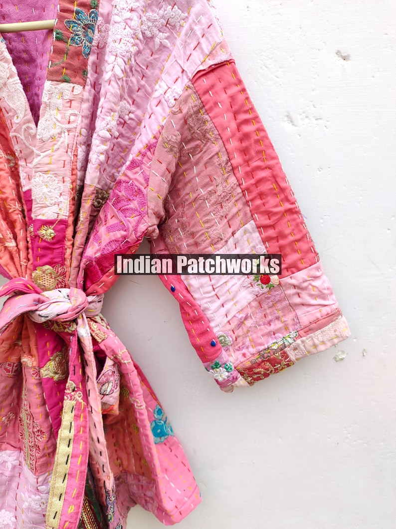 May include: A pink and white patchwork kimono with a tie belt. The kimono is made of different fabrics and has a variety of embroidery details. The text "Indian Patchworks" is visible on the kimono.