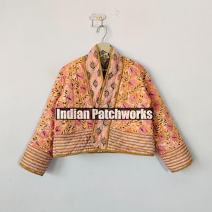 Może przedstawiać: Kurtka pikowana w różowo-pomarańczowym wzorze kwiatowym z kołnierzem mandaryńskim i długimi rękawami. Kurtka ma ozdobne pasy i jest wykonana z bawełny. Na przodzie kurtki nadrukowany jest tekst "Indian Patchworks".
