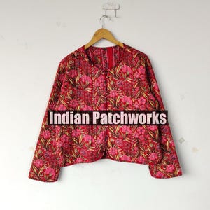 以下が含まれることがあります： ピンクと黄色の花柄の赤いキルティングジャケット。 ジャケットは長袖で、丸いネックラインです。 ジャケットの前面には「Indian Patchworks」の文字が表示されています。