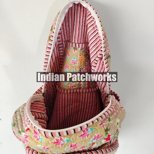 May include: A quilted backpack with a floral pattern in shades of pink, beige, and green. The interior features pink and white striped fabric. The bag has a white zipper and a handle at the top. The text "Indian Patchworks" is visible.