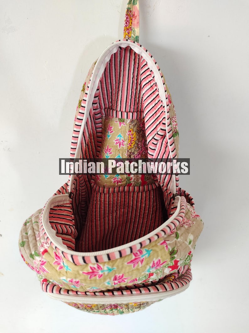 May include: A quilted backpack with a floral pattern in shades of pink, beige, and green. The interior features pink and white striped fabric. The bag has a white zipper and a handle at the top. The text "Indian Patchworks" is visible.