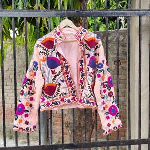 May include: A pink embroidered jacket with floral and geometric patterns in shades of blue, purple, orange, and red. The jacket has a collar and long sleeves.