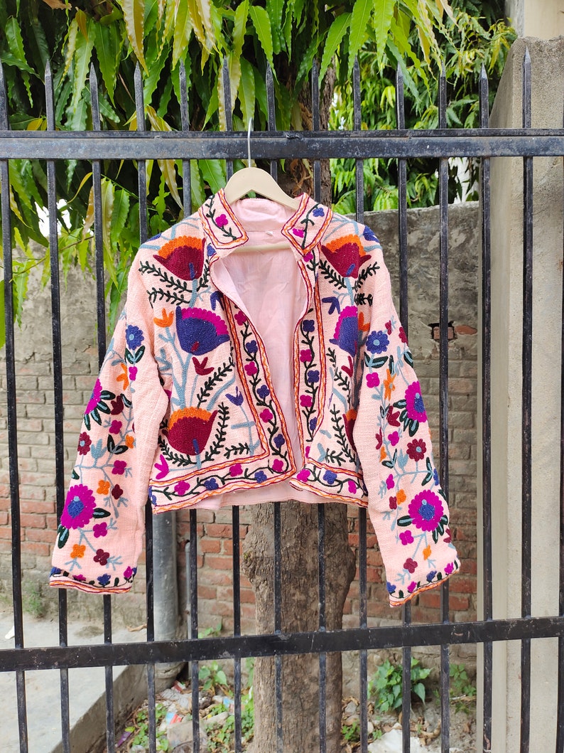 May include: A pink embroidered jacket with floral and geometric patterns in shades of blue, purple, orange, and red. The jacket has a collar and long sleeves.