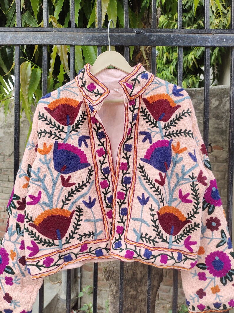 May include: A pink embroidered jacket with a floral pattern. The jacket has a mandarin collar and long sleeves. The embroidery is in shades of blue, green, orange, and purple.
