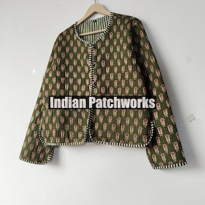 May include: Olive green quilted jacket with a repeating floral pattern in red and cream. The jacket has a striped lining and trim, and the text "Indian Patchworks" is visible. The jacket is hanging on a wooden hanger.