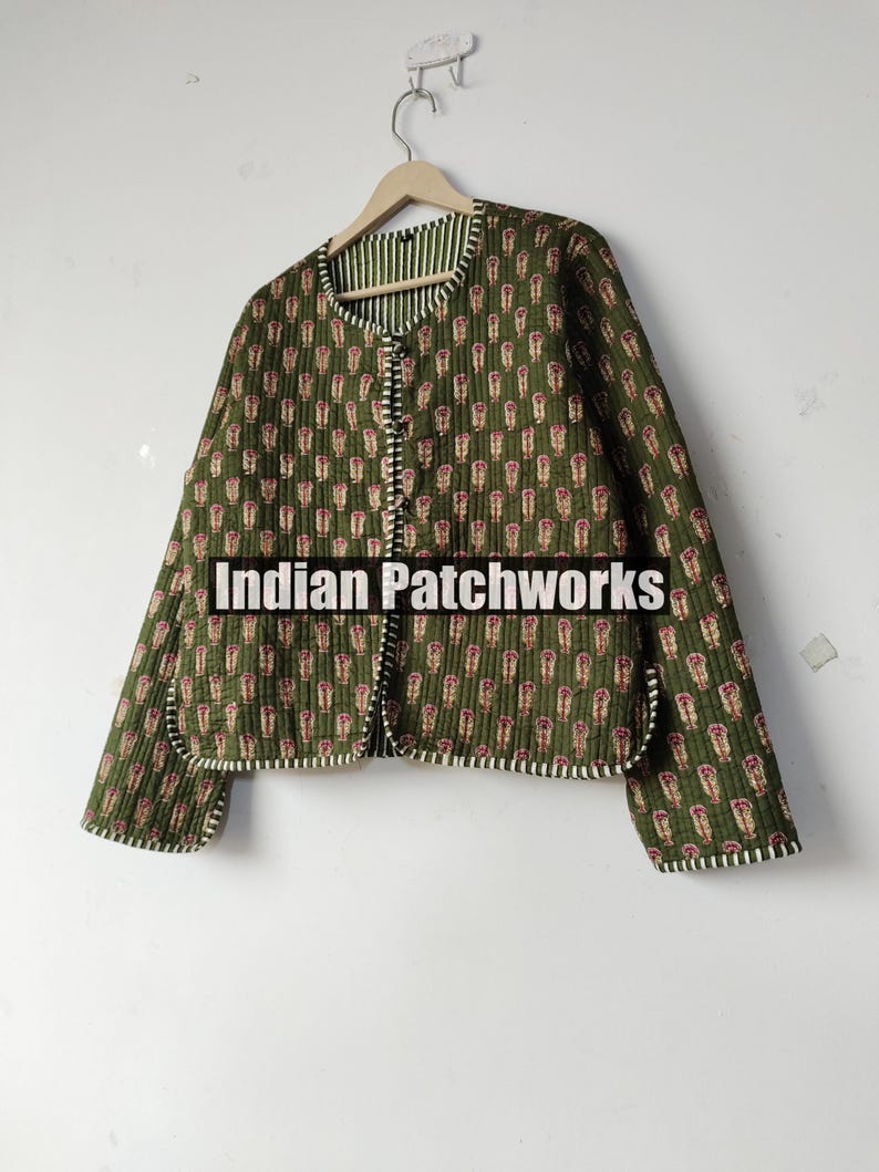 May include: Olive green quilted jacket with a repeating floral pattern in red and cream. The jacket has a striped lining and trim, and the text "Indian Patchworks" is visible. The jacket is hanging on a wooden hanger.