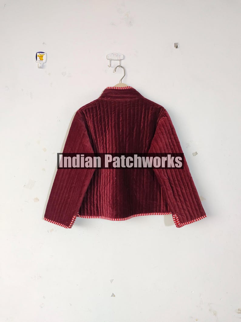 Może przedstawiać: Bordowa pikowana kurtka z wysokim kołnierzem i długimi rękawami. Kurtka ma pionowy wz&oacute;r szw&oacute;w i czerwono-białe paski. Tekst "Indian Patchworks" znajduje się na środku kurtki.