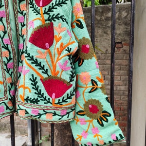 May include: A mint green jacket with colorful floral embroidery. The jacket has a mandarin collar and long sleeves. The embroidery is detailed and intricate, featuring flowers, leaves, and vines.