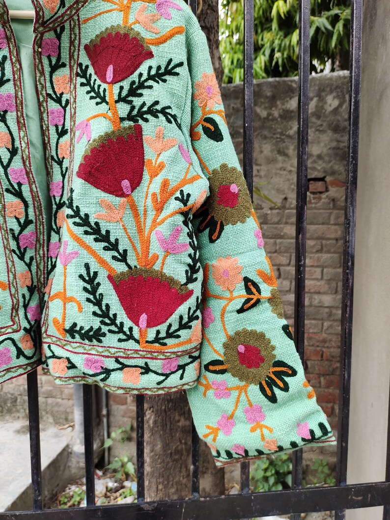May include: A mint green jacket with colorful floral embroidery. The jacket has a mandarin collar and long sleeves. The embroidery is detailed and intricate, featuring flowers, leaves, and vines.