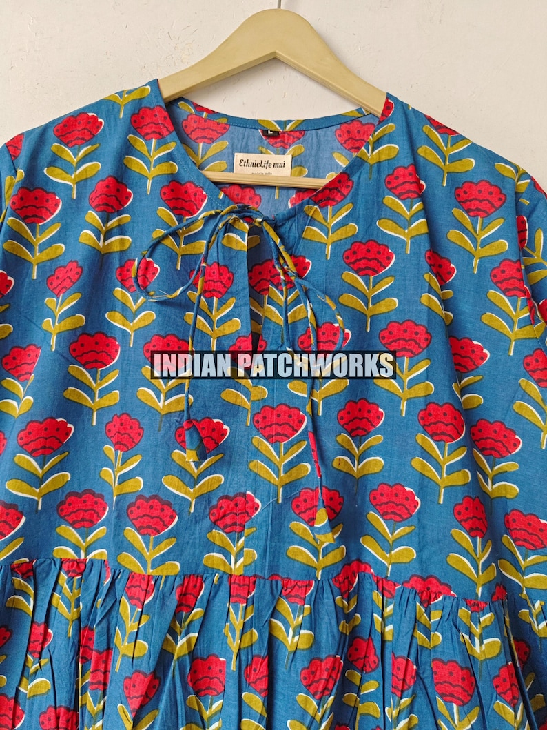 May include: A blue floral print dress featuring red flowers and yellow stems. The dress has a round neckline with a tie closure and a gathered skirt. The text "INDIAN PATCHWORKS" is visible.
