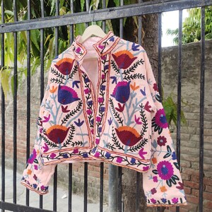 May include: A pink embroidered jacket featuring floral and bird designs in shades of blue, purple, orange, and red. The jacket has a stand-up collar and long sleeves.