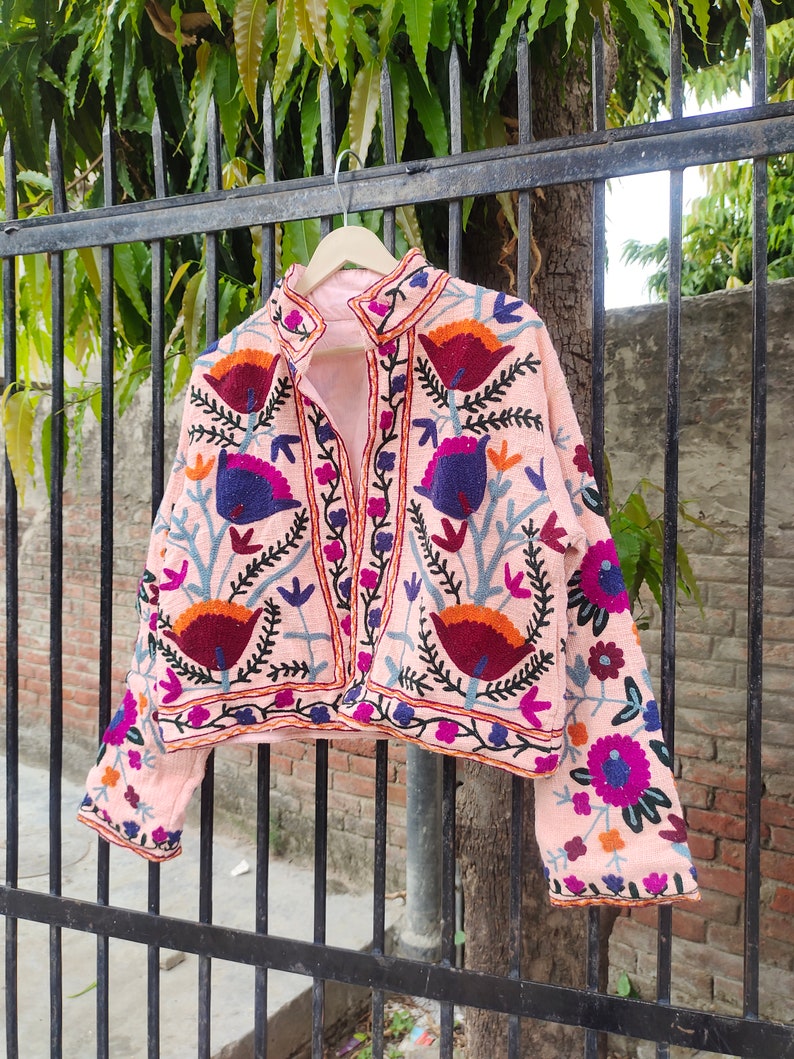 May include: A pink embroidered jacket featuring floral and bird designs in shades of blue, purple, orange, and red. The jacket has a stand-up collar and long sleeves.