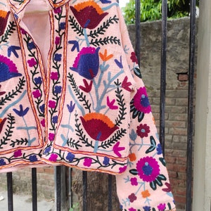 May include: A pink embroidered jacket featuring floral patterns in shades of blue, purple, orange, and red. The jacket has a mandarin collar and a front button closure.