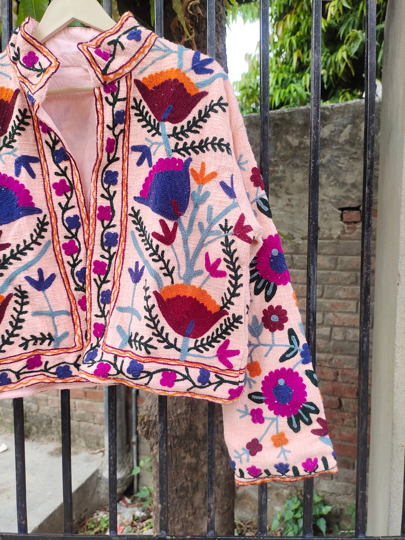 May include: A pink embroidered jacket featuring floral patterns in shades of blue, purple, orange, and red. The jacket has a mandarin collar and a front button closure.