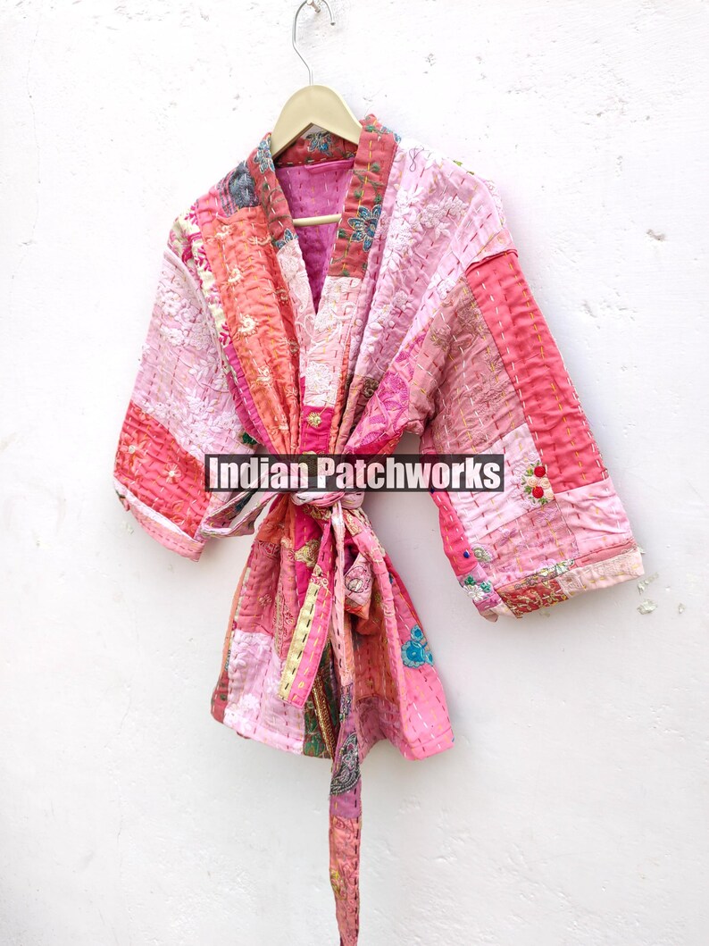 May include: A pink and peach patchwork kimono with a tie belt. The kimono is made of quilted fabric and has a floral design. The text "Indian Patchworks" is visible on the front of the kimono.