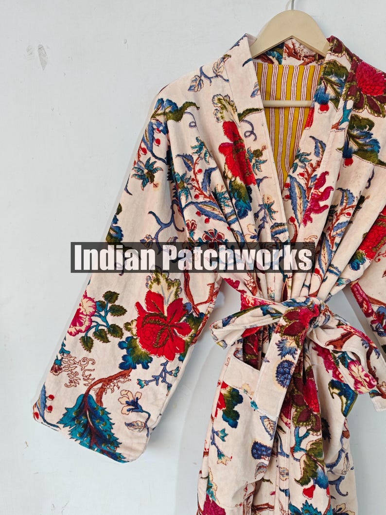May include: A white robe with a floral print in shades of red, blue, and green. The robe has a tie belt and a pocket on the left side. The robe is lined with a yellow and white striped fabric. The text "Indian Patchworks" is visible on the robe.