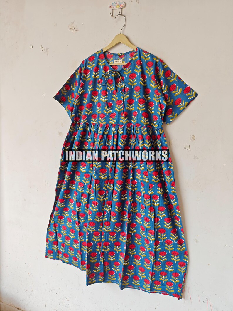 May include: A blue dress with a red floral pattern hangs on a wooden hanger. The dress features short sleeves and a gathered waist. The text "INDIAN PATCHWORKS" is visible. The dress is made of a lightweight fabric, suitable for warmer weather.