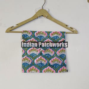 May include: A folded textile with a repeating floral pattern in blue, pink, and green hangs from a wooden hanger. The textile features a symmetrical design of stylized flowers. The text "Indian Patchworks" is across the front of the textile.