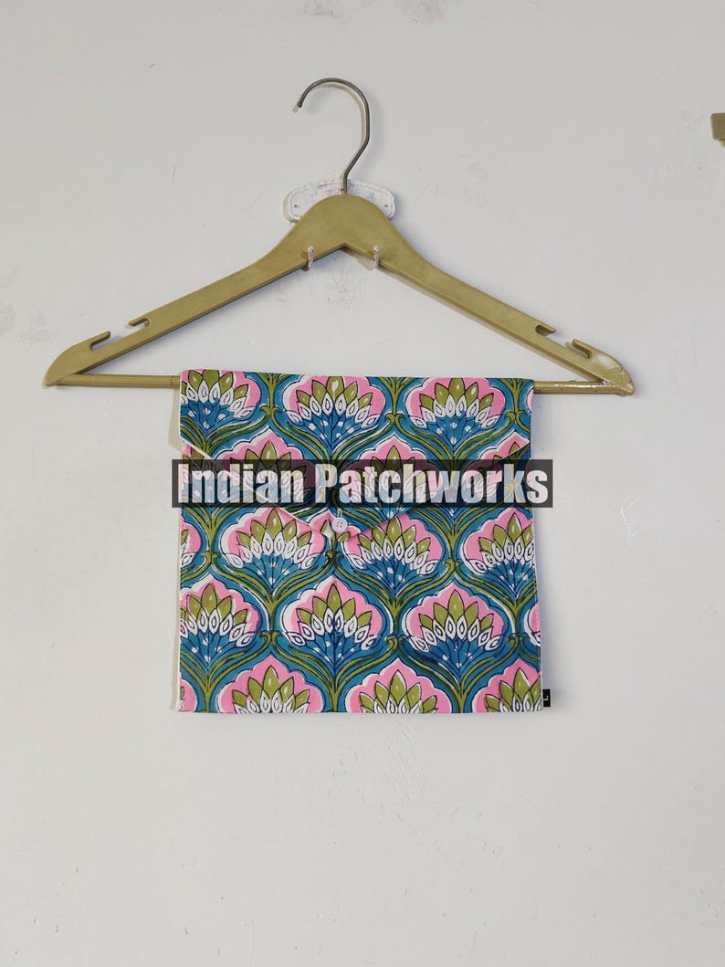 May include: A folded textile with a repeating floral pattern in blue, pink, and green hangs from a wooden hanger. The textile features a symmetrical design of stylized flowers. The text "Indian Patchworks" is across the front of the textile.