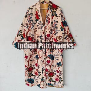 May include: A long, cream-colored robe with a floral print in shades of red, blue, and green. The robe has a yellow and white striped lining and is hanging on a hanger. The text "Indian Patchworks" is visible on the front of the robe.