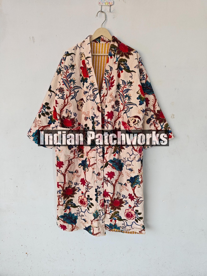 May include: A long, cream-colored robe with a floral print in shades of red, blue, and green. The robe has a yellow and white striped lining and is hanging on a hanger. The text "Indian Patchworks" is visible on the front of the robe.