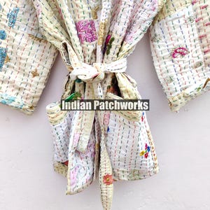 K&ouml;nnte beinhalten: Eine wei&szlig;e Baumwollrobe mit bunten Stickereien und Patchwork-Details. Die Robe hat einen Bindeg&uuml;rtel und zwei Taschen. Der Text "Indian Patchworks" ist auf der Robe sichtbar.