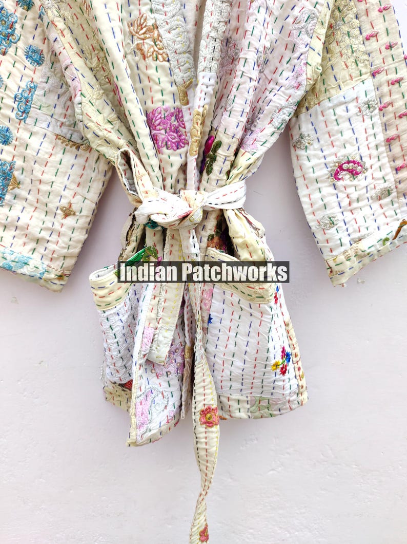 K&ouml;nnte beinhalten: Eine wei&szlig;e Baumwollrobe mit bunten Stickereien und Patchwork-Details. Die Robe hat einen Bindeg&uuml;rtel und zwei Taschen. Der Text "Indian Patchworks" ist auf der Robe sichtbar.