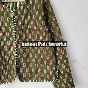 May include: Olive green quilted jacket with a repeating floral pattern in pink and beige. The jacket has a striped lining and trim. The text "Indian Patchworks" is displayed on the bottom of the image. The jacket is buttoned up.