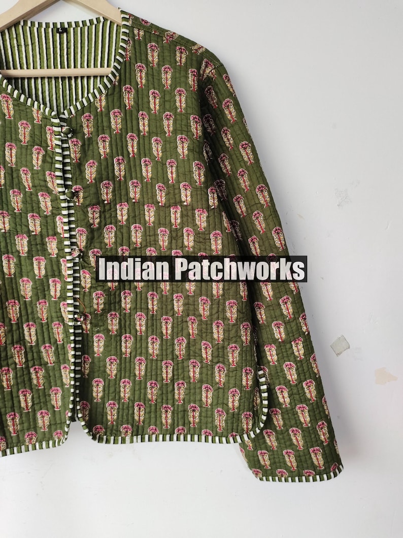May include: Olive green quilted jacket with a repeating floral pattern in pink and beige. The jacket has a striped lining and trim. The text "Indian Patchworks" is displayed on the bottom of the image. The jacket is buttoned up.