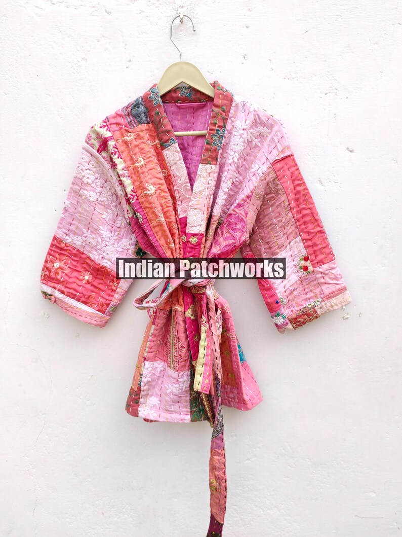 May include: A pink and red patchwork quilted kimono with a long tie belt. The kimono has a V-neck and is made of a soft, lightweight fabric. "Indian Patchworks" is printed on the front of the kimono.