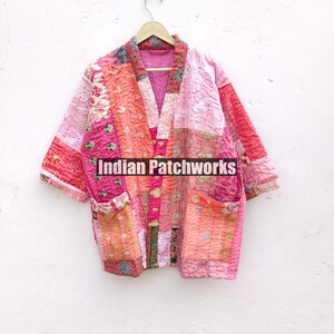 May include: A pink and orange patchwork quilted jacket with a V-neck and pockets. The jacket is hanging on a wooden hanger against a white wall. The text "Indian Patchworks" is visible on the jacket.