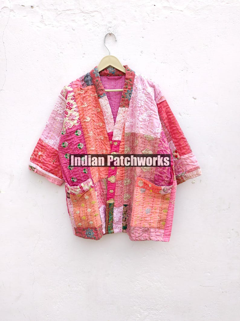 May include: A pink and orange patchwork quilted jacket with a V-neck and pockets. The jacket is hanging on a wooden hanger against a white wall. The text "Indian Patchworks" is visible on the jacket.