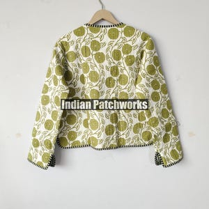 May include: A quilted jacket with a white background and a repeating pattern of green floral designs. The jacket has a black and white trim along the collar and cuffs. The jacket is hanging on a wooden hanger.