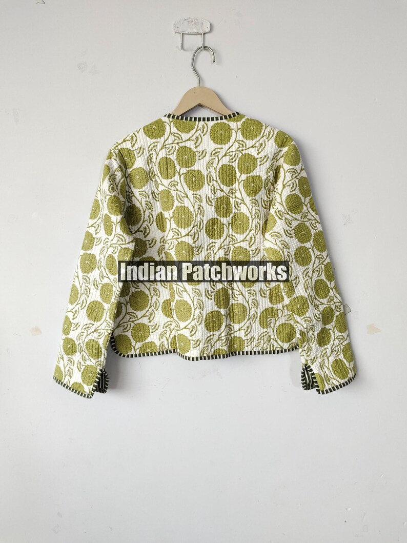 May include: A quilted jacket with a white background and a repeating pattern of green floral designs. The jacket has a black and white trim along the collar and cuffs. The jacket is hanging on a wooden hanger.