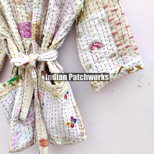 K&ouml;nnte beinhalten: Eine wei&szlig;e Baumwollrobe mit bunten Stickereien und Patchwork-Details. Die Robe hat einen Bindeg&uuml;rtel und Taschen. Der Text "Indian Patchworks" ist auf der Robe sichtbar.