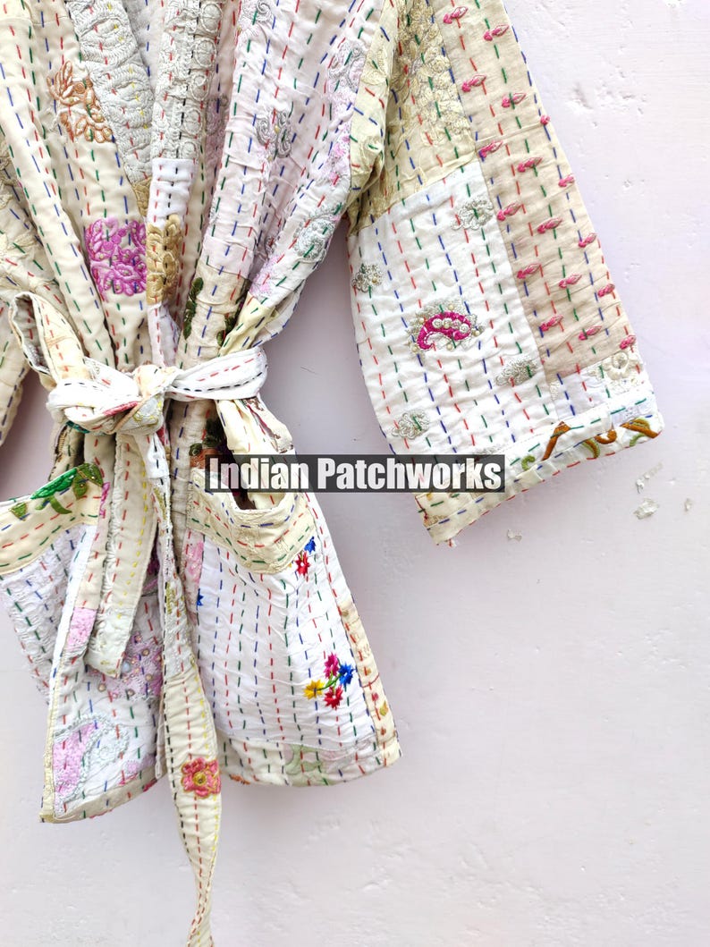 K&ouml;nnte beinhalten: Eine wei&szlig;e Baumwollrobe mit bunten Stickereien und Patchwork-Details. Die Robe hat einen Bindeg&uuml;rtel und Taschen. Der Text "Indian Patchworks" ist auf der Robe sichtbar.