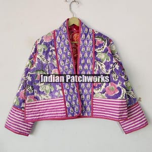 Peut inclure: Une veste matelassée violette et rose avec un imprimé floral et une doublure rose. La veste a un col mandarin et une longueur courte. Le texte "Indian Patchworks" est visible sur la veste.