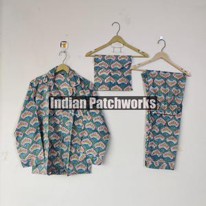 May include: A three-piece pajama set with a floral pattern in shades of teal, pink, and white. The set includes a button-down shirt, a pair of pants, and a matching eye mask, all hanging on wooden hangers. The text "Indian Patchworks" is visible.