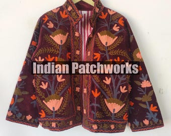 Op de afbeelding: Een donkerrode fluwelen jas met bloemenborduurwerk in tinten oranje, roze, groen en blauw. De jas heeft een mandarijnsluiting en een knoopsluiting. De tekst "Indian Patchworks" is zichtbaar op de voorkant van de jas.