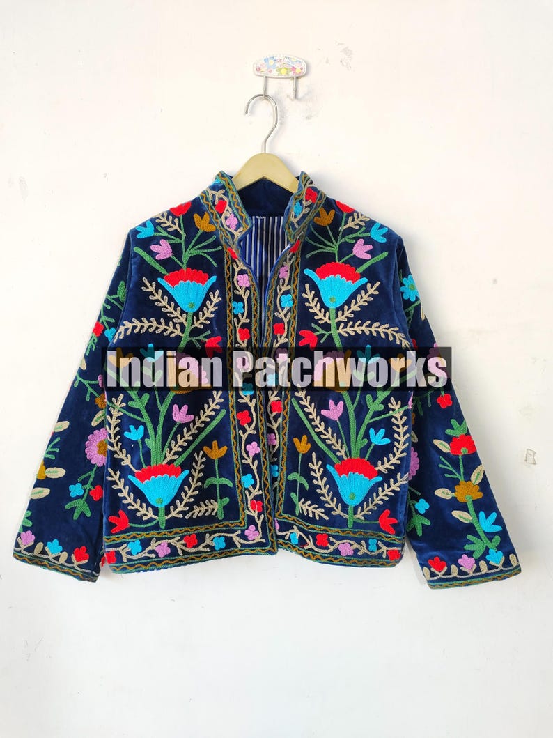 May include: A navy blue velvet jacket with colorful floral embroidery. The jacket has a mandarin collar and a zipper closure. The embroidery is intricate and detailed, featuring a variety of flowers and leaves. The text "Indian Patchworks" is visible on the jacket.