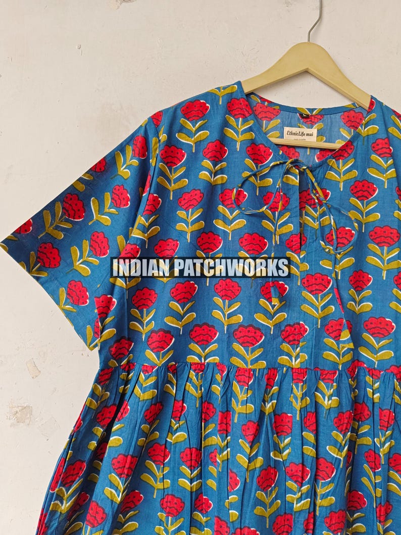 May include: A blue dress featuring a repeating pattern of red flowers with yellow stems. The dress has short sleeves and a tie at the neckline. The text "INDIAN PATCHWORKS" is across the front.
