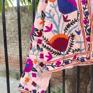 May include: A pink embroidered jacket featuring floral patterns in shades of blue, orange, red, and purple. The jacket has a cropped length and a slightly flared silhouette.