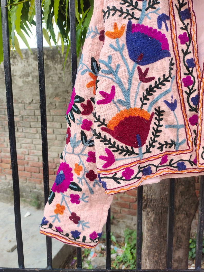 May include: A pink embroidered jacket featuring floral patterns in shades of blue, orange, red, and purple. The jacket has a cropped length and a slightly flared silhouette.