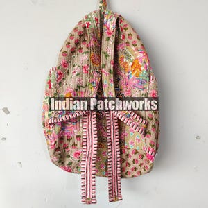 May include: A floral patterned backpack in shades of pink, green, and yellow, with a beige background. The backpack has pink and white striped straps and is hanging on a white wall. The text "Indian Patchworks" is across the front.