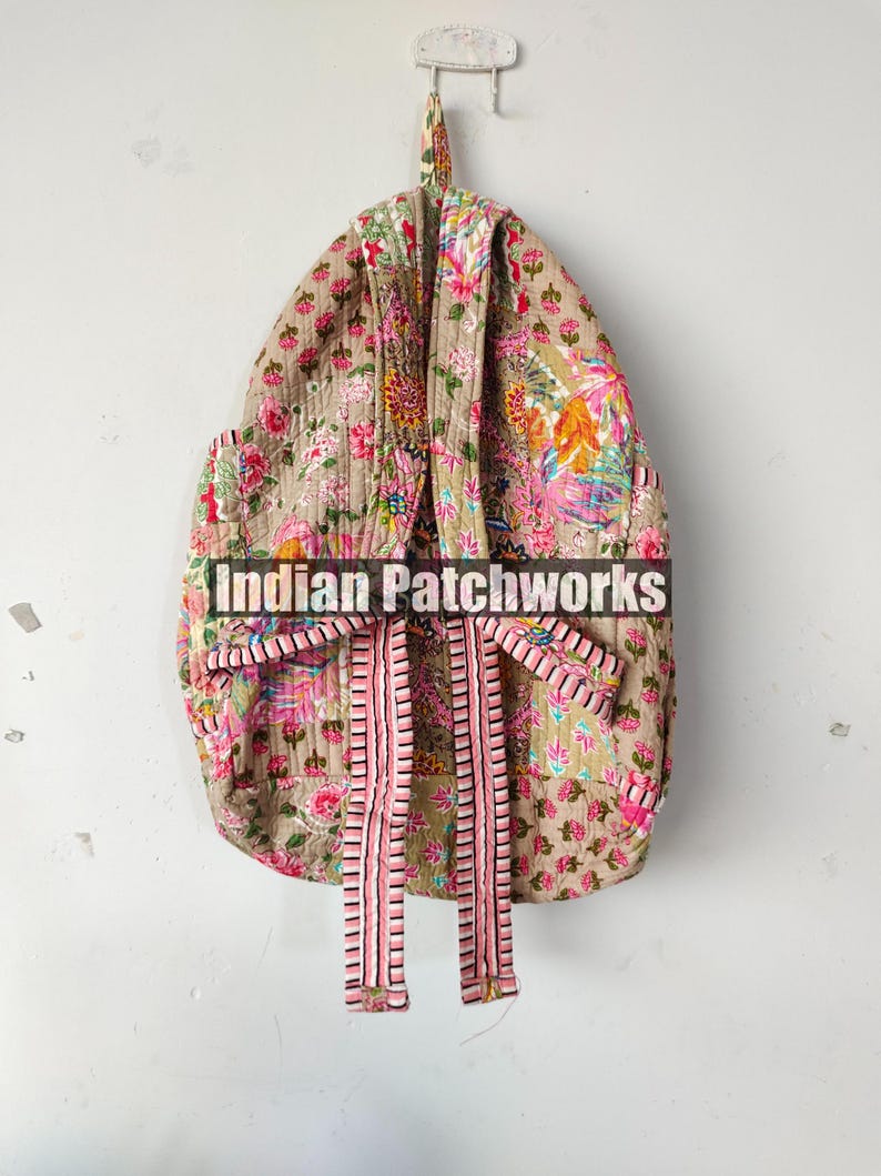 May include: A floral patterned backpack in shades of pink, green, and yellow, with a beige background. The backpack has pink and white striped straps and is hanging on a white wall. The text "Indian Patchworks" is across the front.