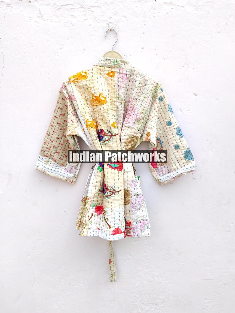 K&ouml;nnte beinhalten: Eine wei&szlig;e Baumwollrobe mit einem floralen Patchwork-Muster. Die Robe hat einen Bindeg&uuml;rtel und lange &Auml;rmel. Der Text "Indian Patchworks" ist auf der Robe sichtbar.