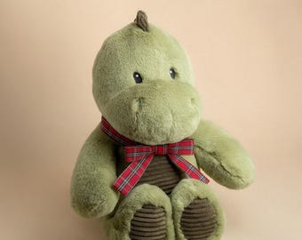 May include: A plush dinosaur toy named Dash the Dinosaur by Furrful, featuring soft sage green fur, a dark brown corduroy belly and foot pads, and a red, green, and white plaid ribbon tied in a bow around its neck. The text Furrful Dash the Dinosaur is visible.