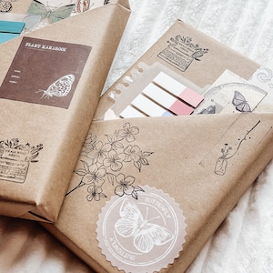 Könnte beinhalten: Zwei in braunes Papier verpackte Pakete mit Vintage-Ästhetik. Ein Paket hat ein Etikett mit der Aufschrift "Plant Handbook" und das andere einen Aufkleber mit der Aufschrift "Butterfly". Beide Pakete haben dekorative Stempel mit Blumen- und Schmetterlingsmotiven.